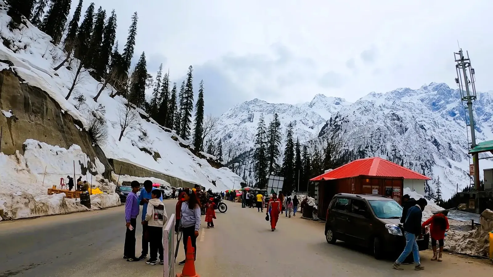 Snow-lined mountain road near Manali with tourists, a red-roofed kiosk and snowy peaks
