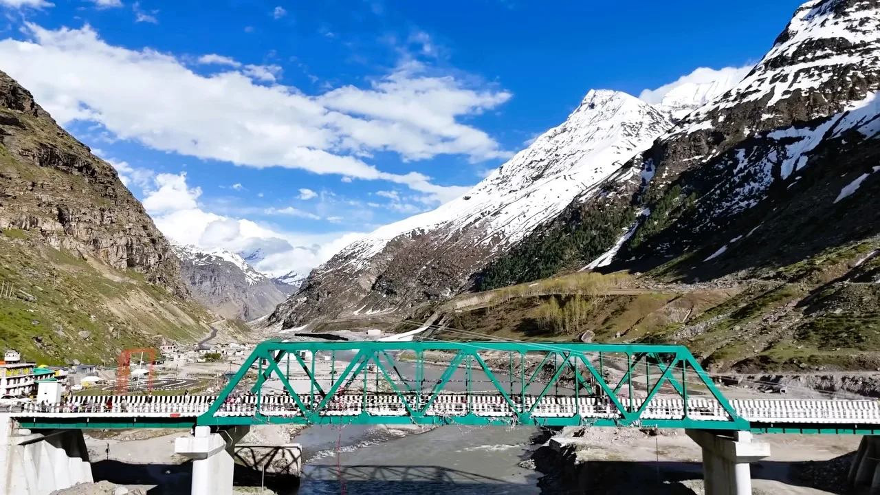 Green steel bridge over a mountain river below snow-capped peaks